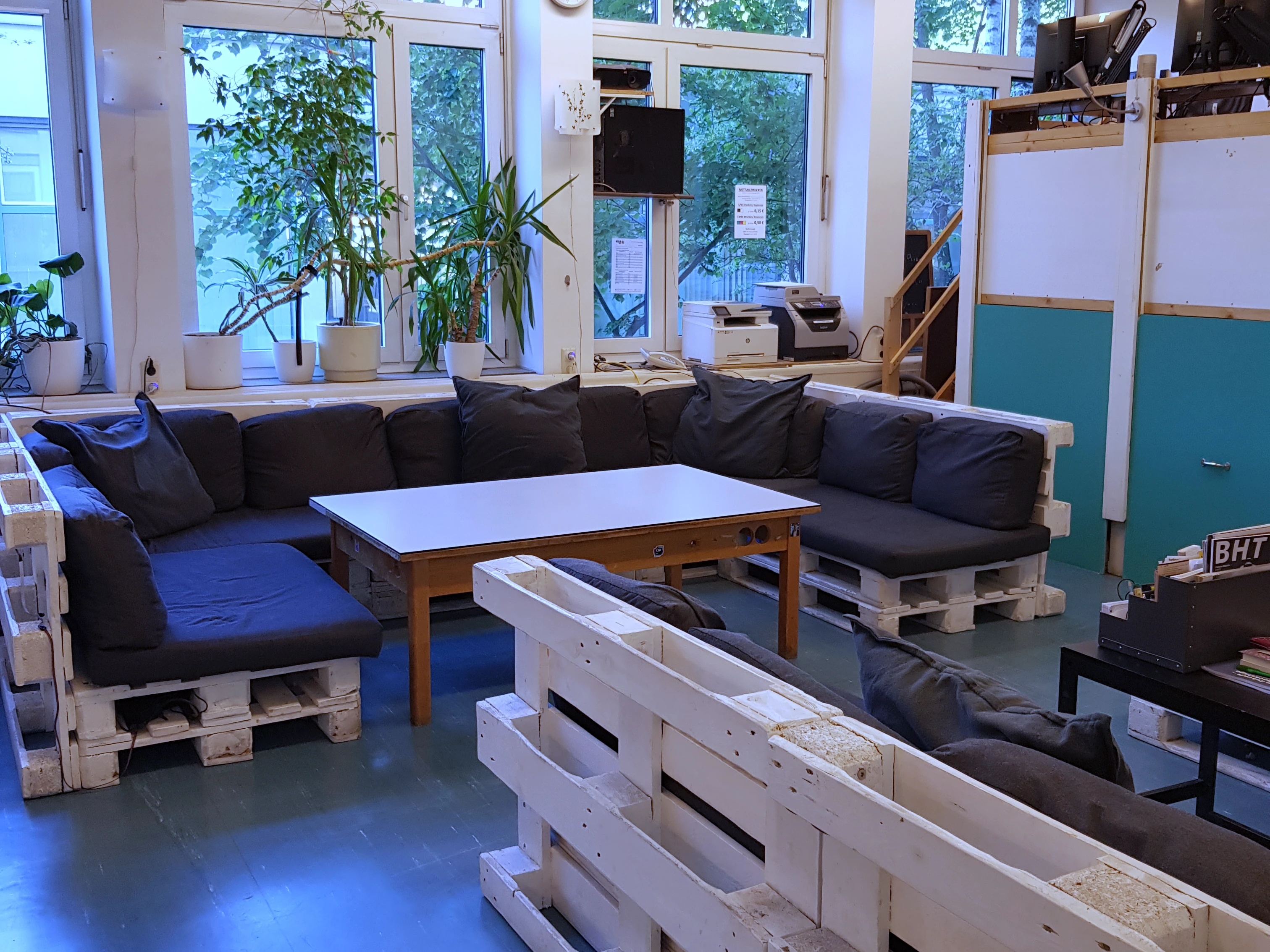 Foto eines Raumes, im Vordergrund sind zwei Gruppen Sofas aus Euro-Paletten zu sehen, im Hintergrund eine Tribüne mit Arbeitsplätzen sowie eine Fensterfront mit Topfpflanzen.